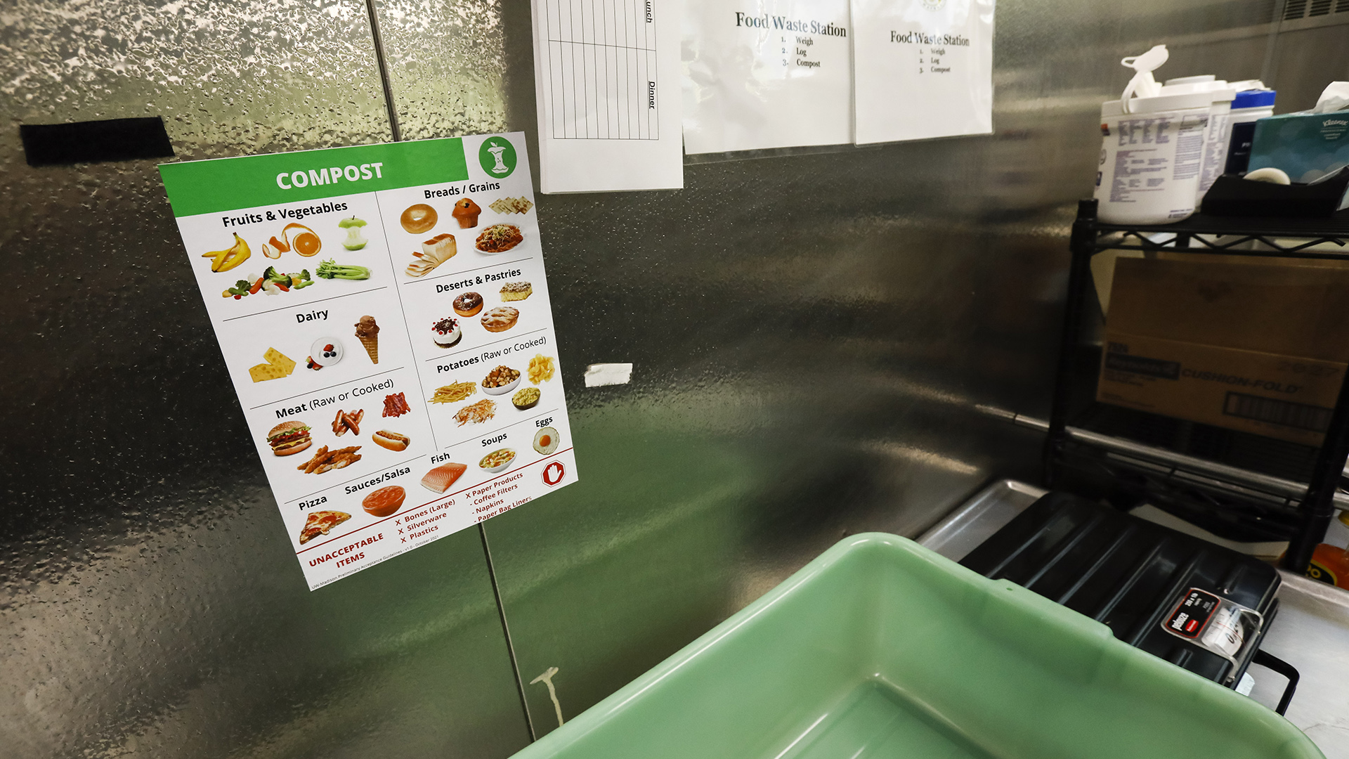 A sign indicating food items that can be composted is attached to a metal wall inside a commercial food preparation space above a plastic tub in an area labeled as a food waste station.