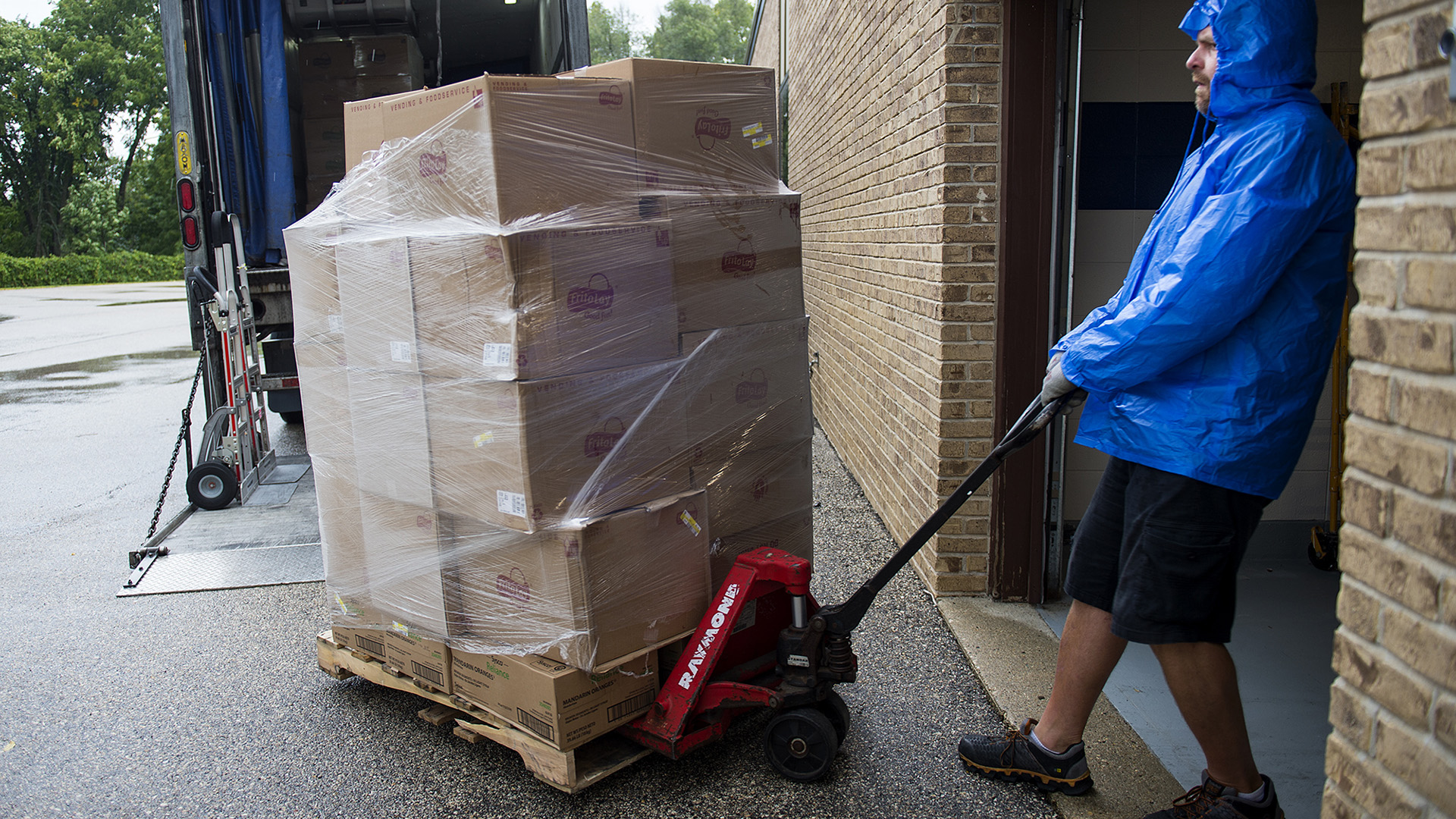 A man wearing a raincoat pulls a hydrualic pallet jack stacked with plastic-wrapped cardboard boxes through a large door facing a driveway in which a truck is parked.