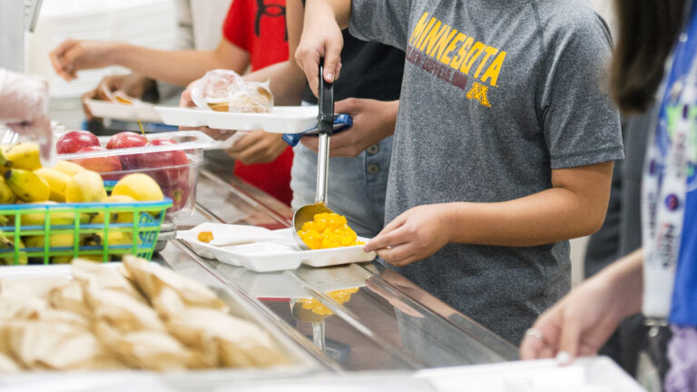Students face a metal shelf for trays select food items from the adjacent counter, with one using a ladle to scoop fruit into a clamshell food container.