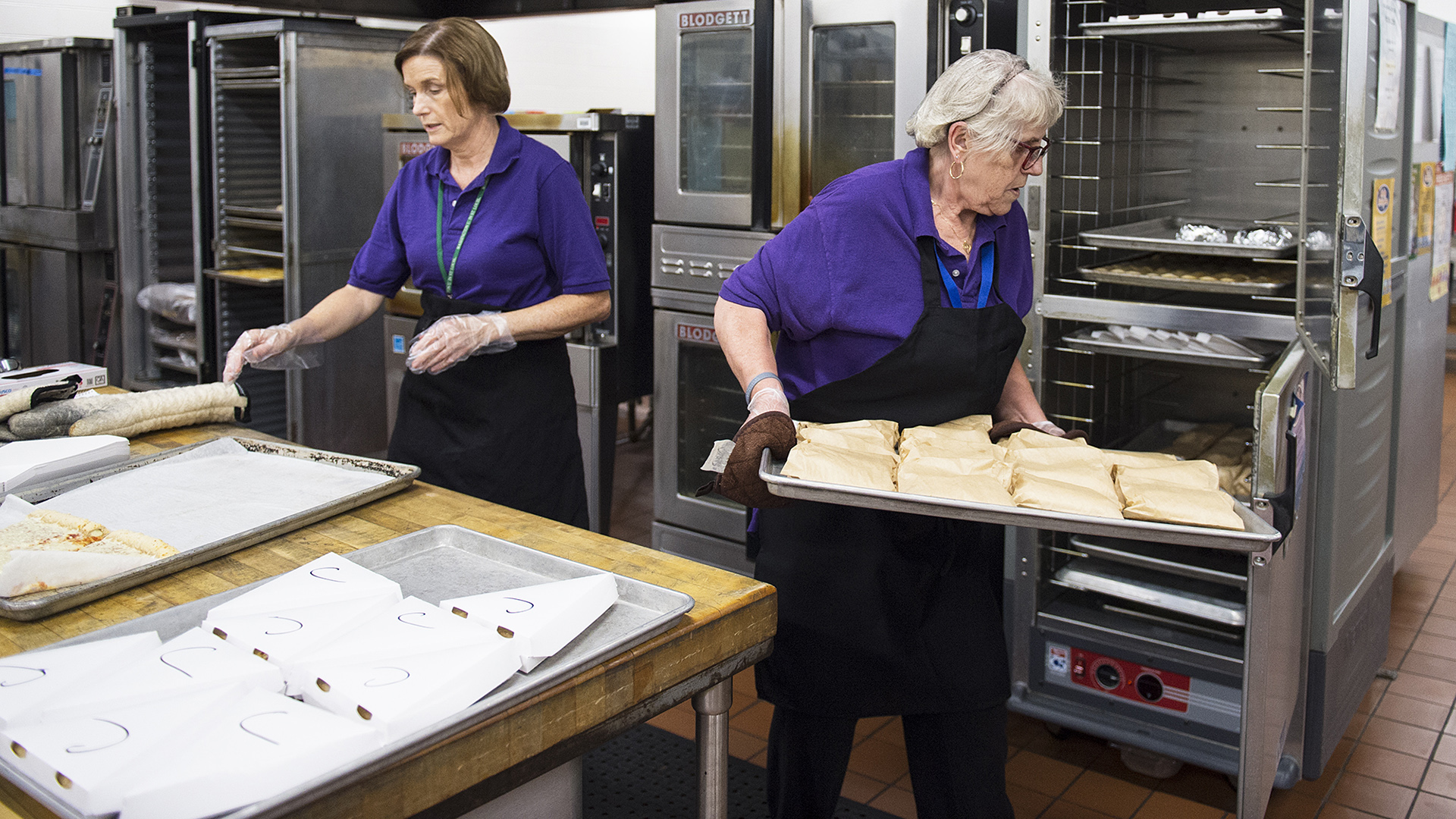 Two people wearing matching shirts and aprons work in a commercial kitchen, with one facing a large table with a butcher's block surface that is covered with large backing trays, and another placing one tray filled with food items wrapped in paper into an adjacent heating appliance, with other institutional food service equipment in the background.