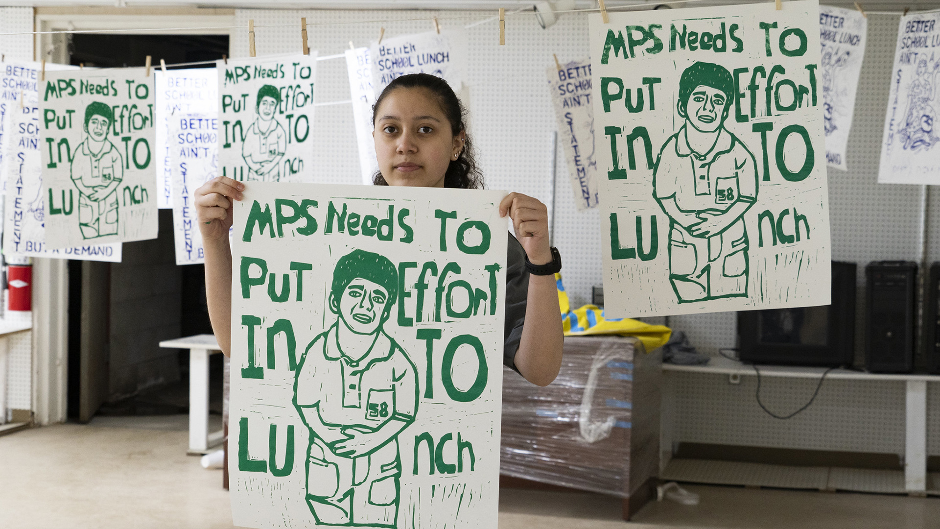 Doricela Herrera-Sanchez poses for a portrait while holding a screen-printed poster with a graphic of a person holding their stomach and the words "MPS Need To Put Effort Into Lunch," with additional posters of this design and others hanging on clotheslines in a room with pegboard walls.