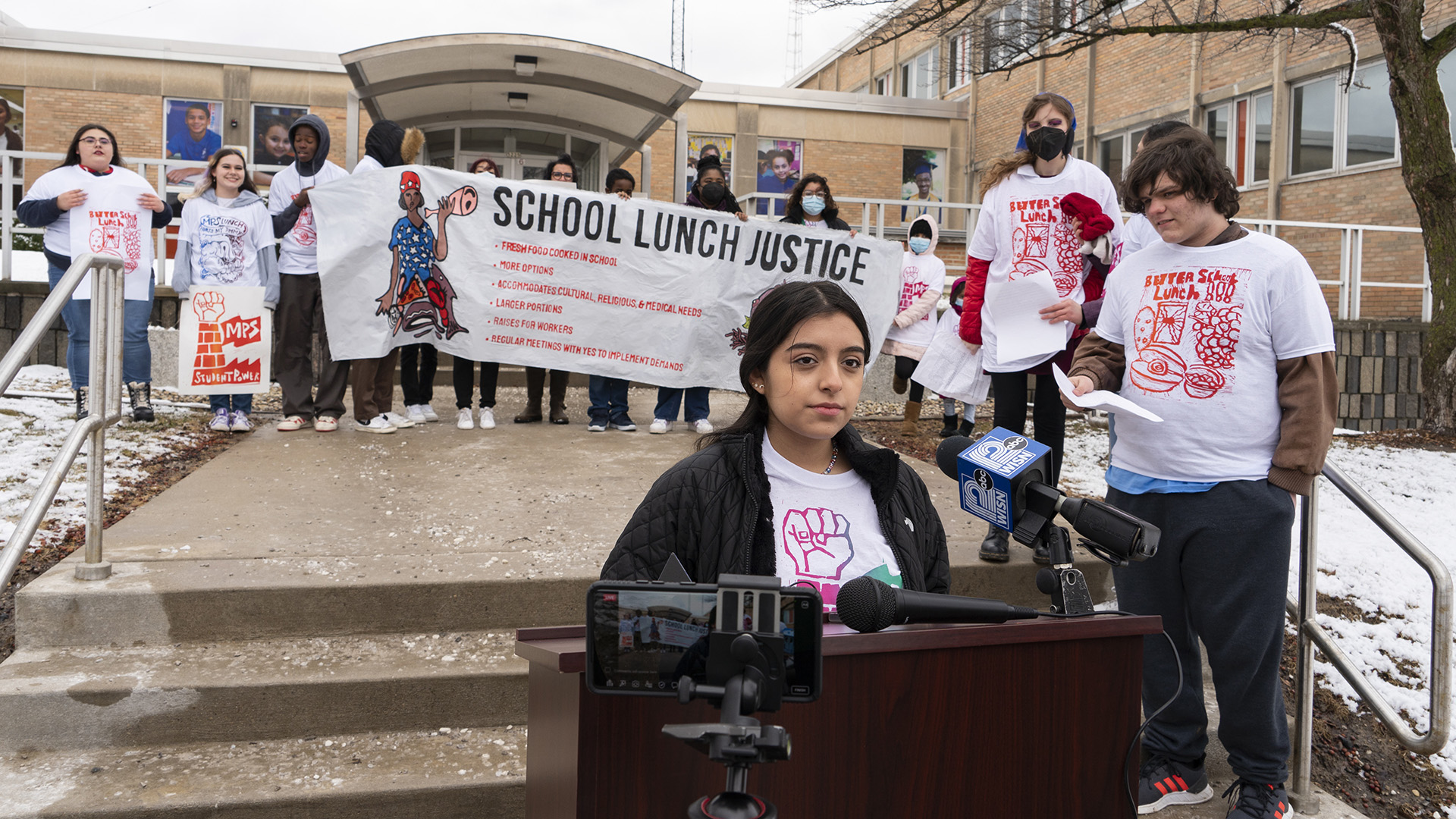 A student stands at a podium with outfitted with two microphones and in front of a mobile phone recording video, and in front of more students standing on the a short concrete staircase and sidewalk who are holding posters and a sign reading "School Lunch Justice," with a school building in the background.
