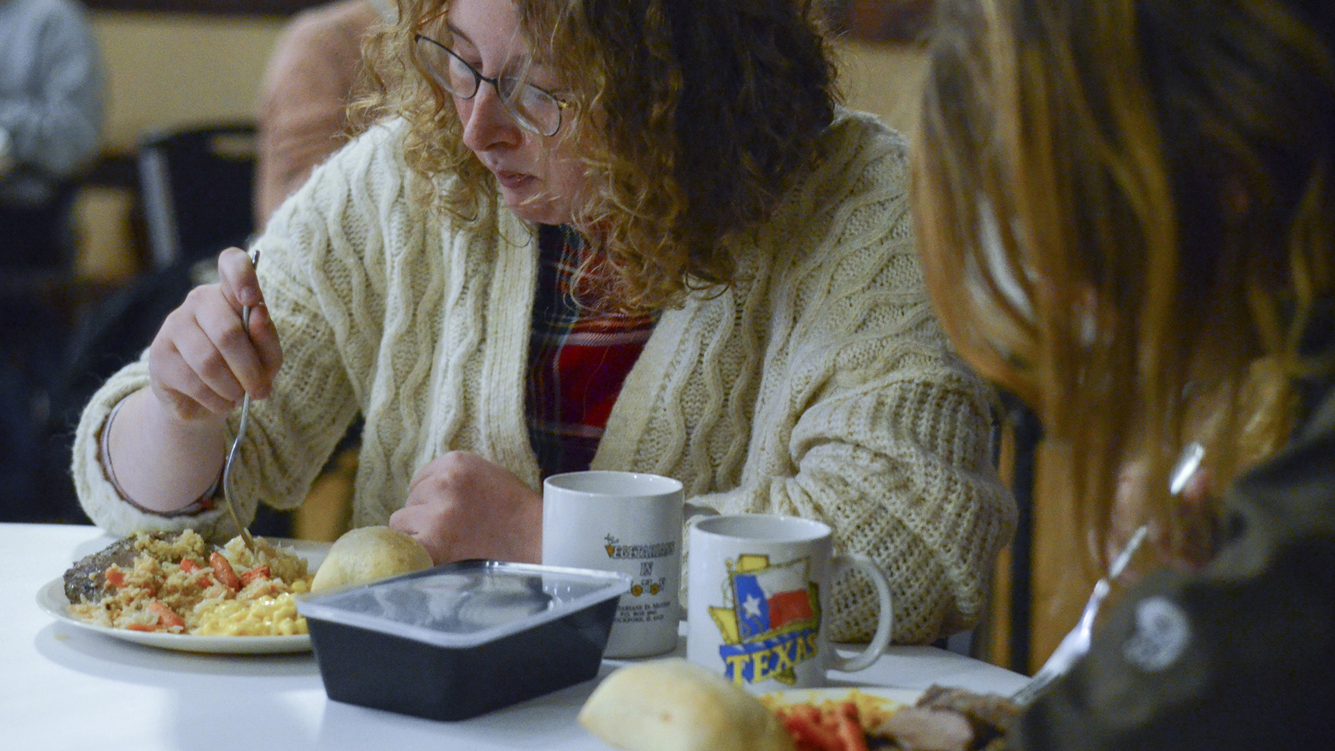 Thea Sutter sits at a table and eats from a food-covered plate that sits on a table next to a disposable to-go container, two coffee mugs and another plate of food, with another person in profile in the foreground.