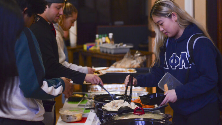 Julia Zhang stands on one side of a buffet and uses tongs with her right hand to select a food item for placing in a disposable container in her left hand, with other people selecting food items from the other side of the table.