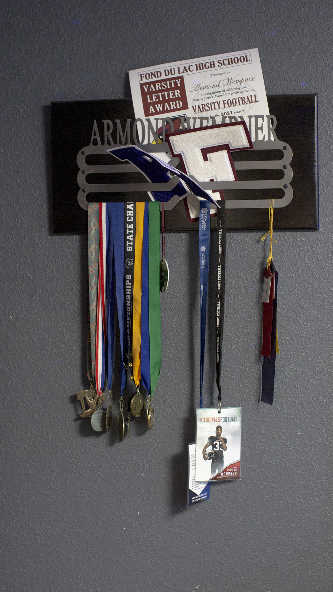 Medals, ID cards on lanyards, high-school letters, ribbons and other items cover a wall-mounted metal and wood display with the name "Armond Wempner."
