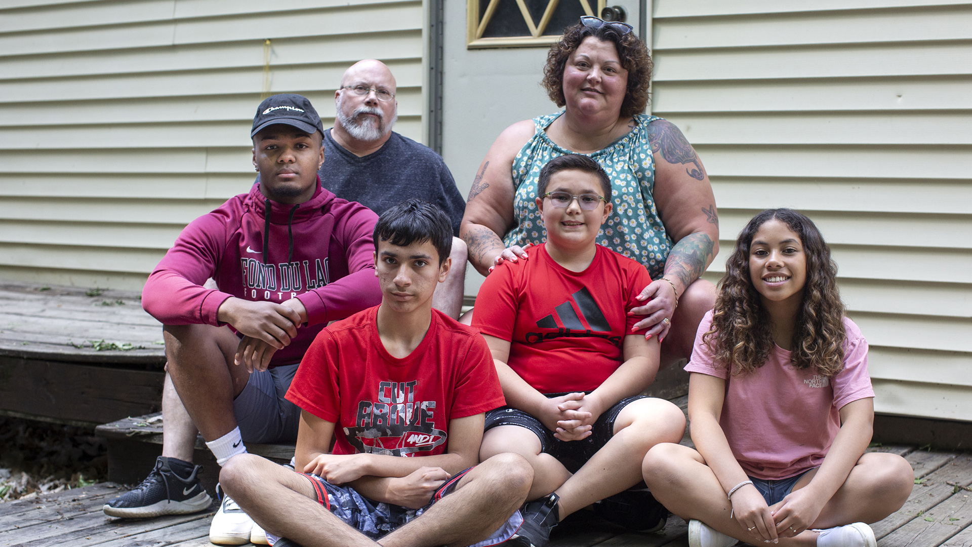 Armond, Dan, Mason, Amy, Cameron and Merri Wempner sit on steps and deck outside the exterior door of a house.