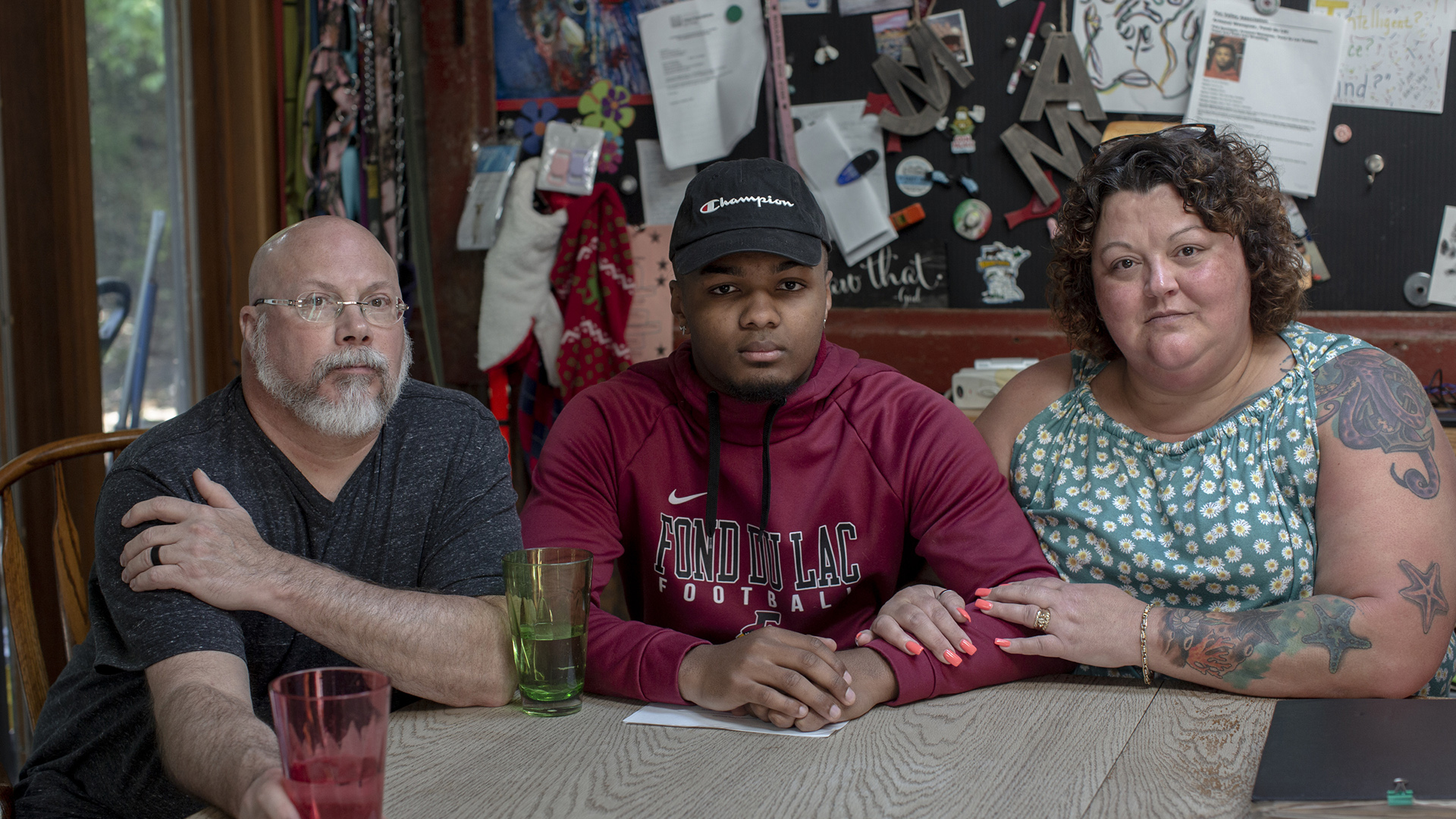 Dan, Armond and Amy Wempner sit at a table, with a window and a bulletin board covered with pins, papers and other items in the background.