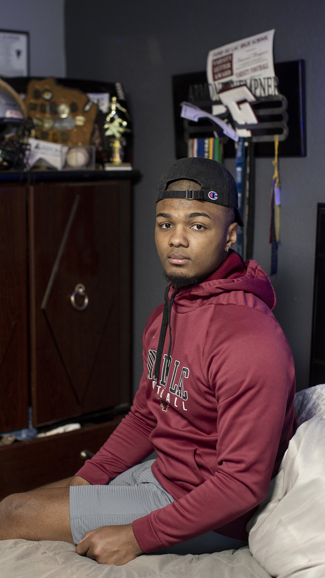 Armond Wempner sits on a bed with an armoire covered with trophies and a wall-mounted rack for medals in the background.
