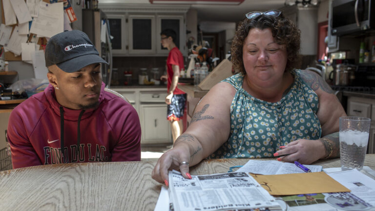 Armond and Amy Wempner sit at a table in a kitchen and look at papers arrayed in front of them, with appliances, cupboards, counters and another person standing in the background.