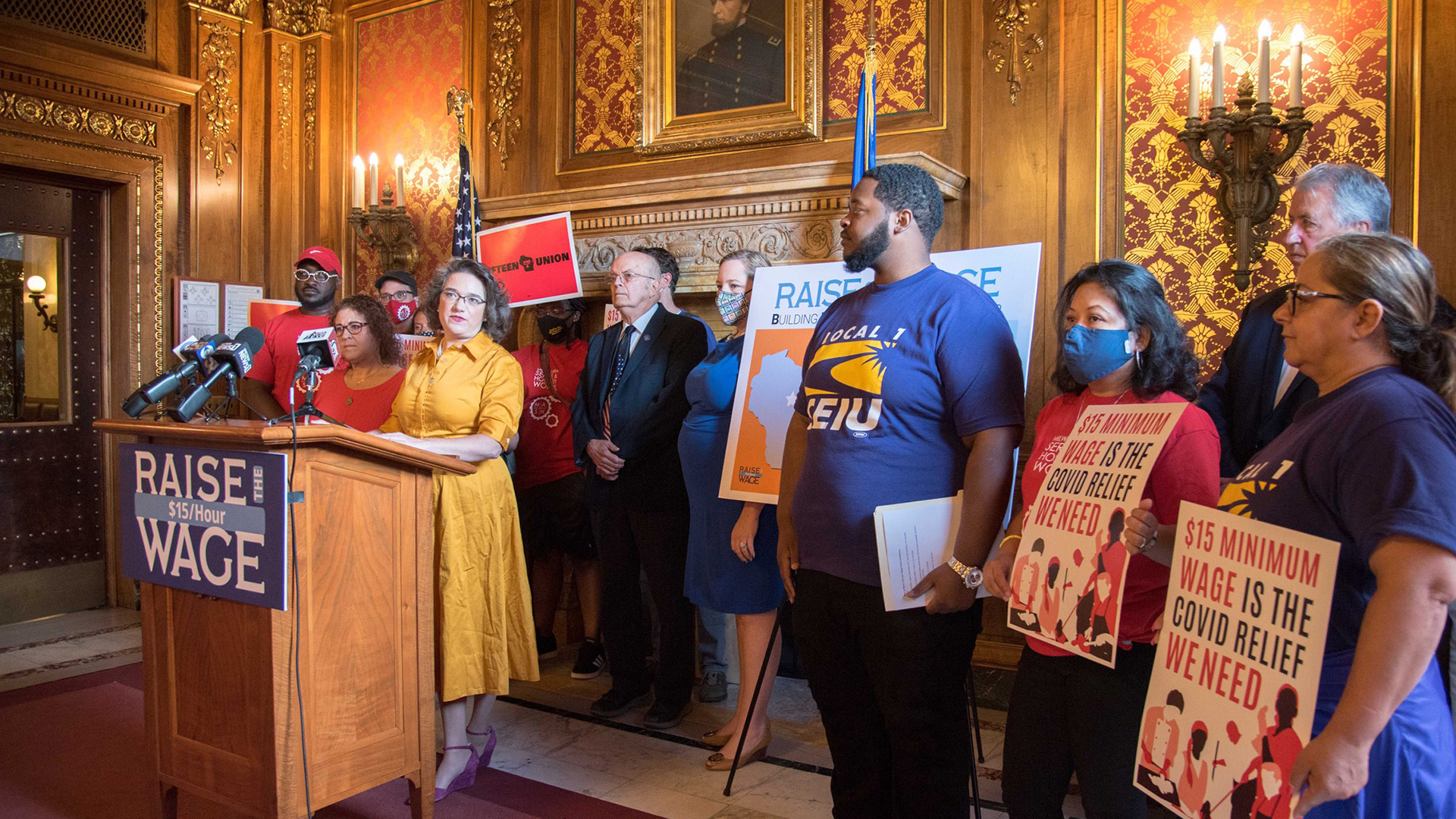 Lisa Subeck and Melissa Agard stand behind a wood podium with multiple microphones and a sign reading "Raise Wage" and "$15/Hour," with other people standing behind and on either side of them, with several holding a poster reading "$15 Minimum Wage is the Covid Relief We Need" in room with ornate wood paneling, wallpaper and wall sconces.