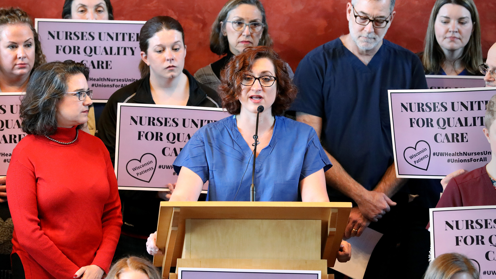 Mariah Clark stands behind a podium and speaks into a microphone, with other people standing on the sides and in the background, with several holding signs that read "Nurses United for Quality Care."