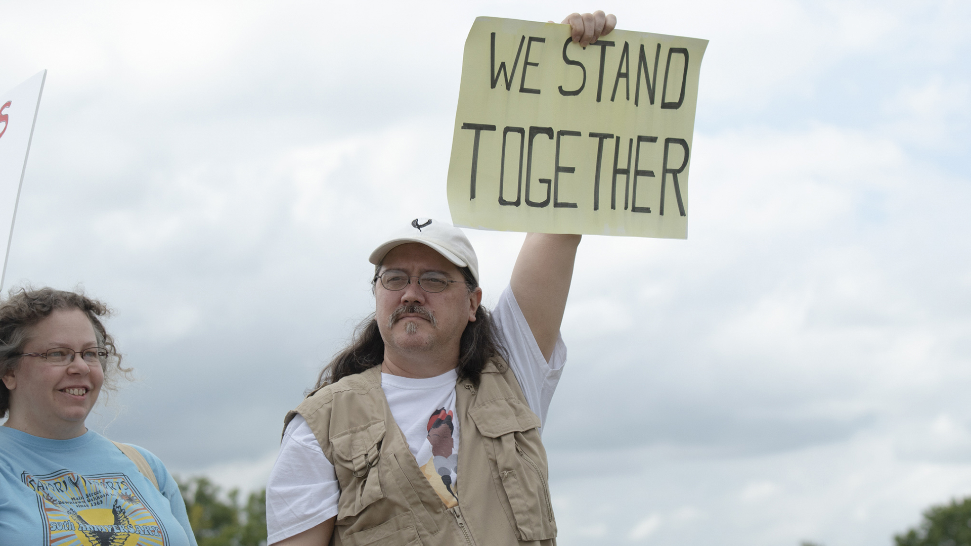 Bob Knudsen holds a hand-drawn sign reading "We Stand Together" above his head while standing next to Lori Knudsen.