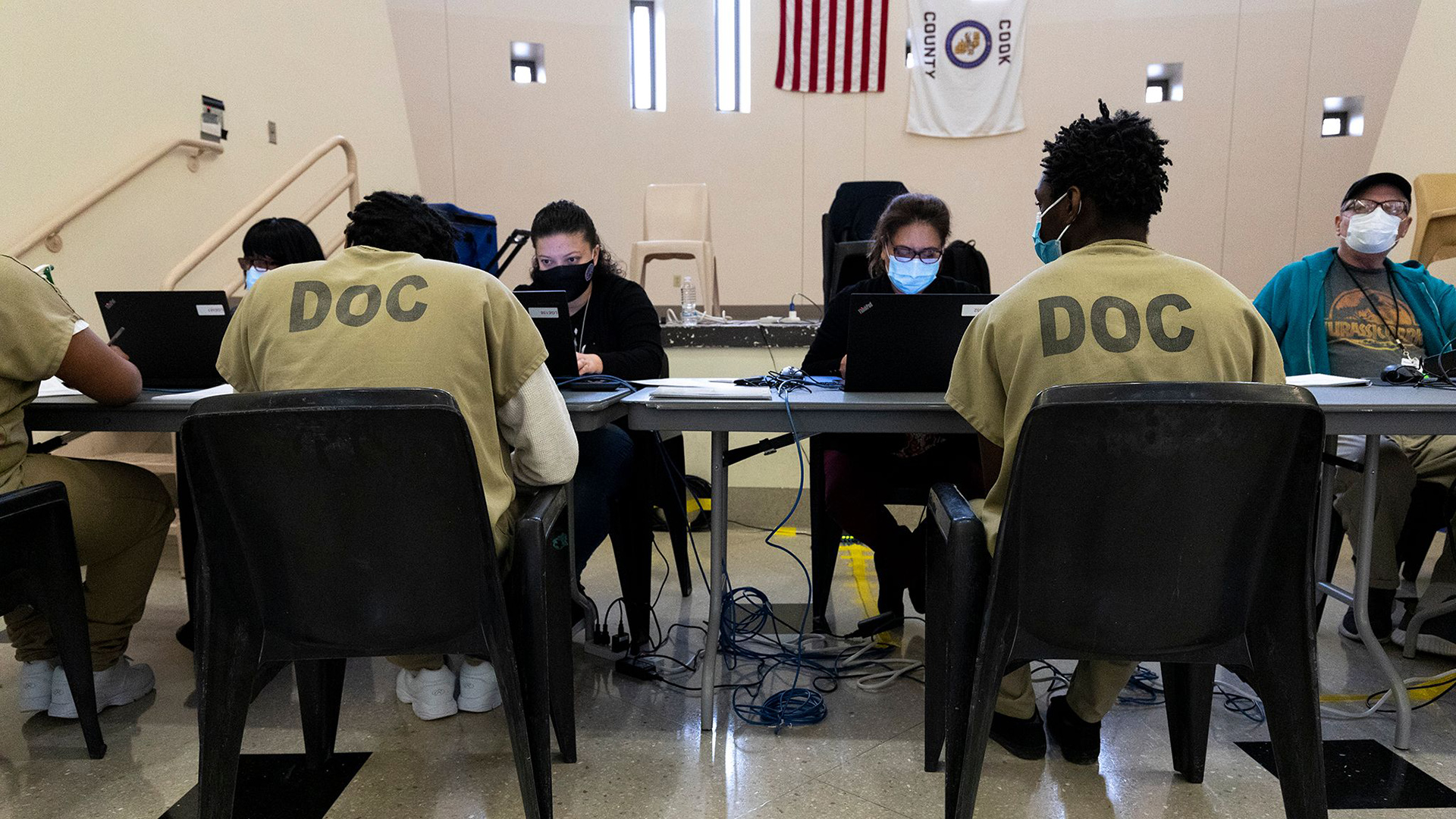 Three inmates sit at a table facing poll workers using laptops to conduct voting in a room with tall narrow and small square windows.