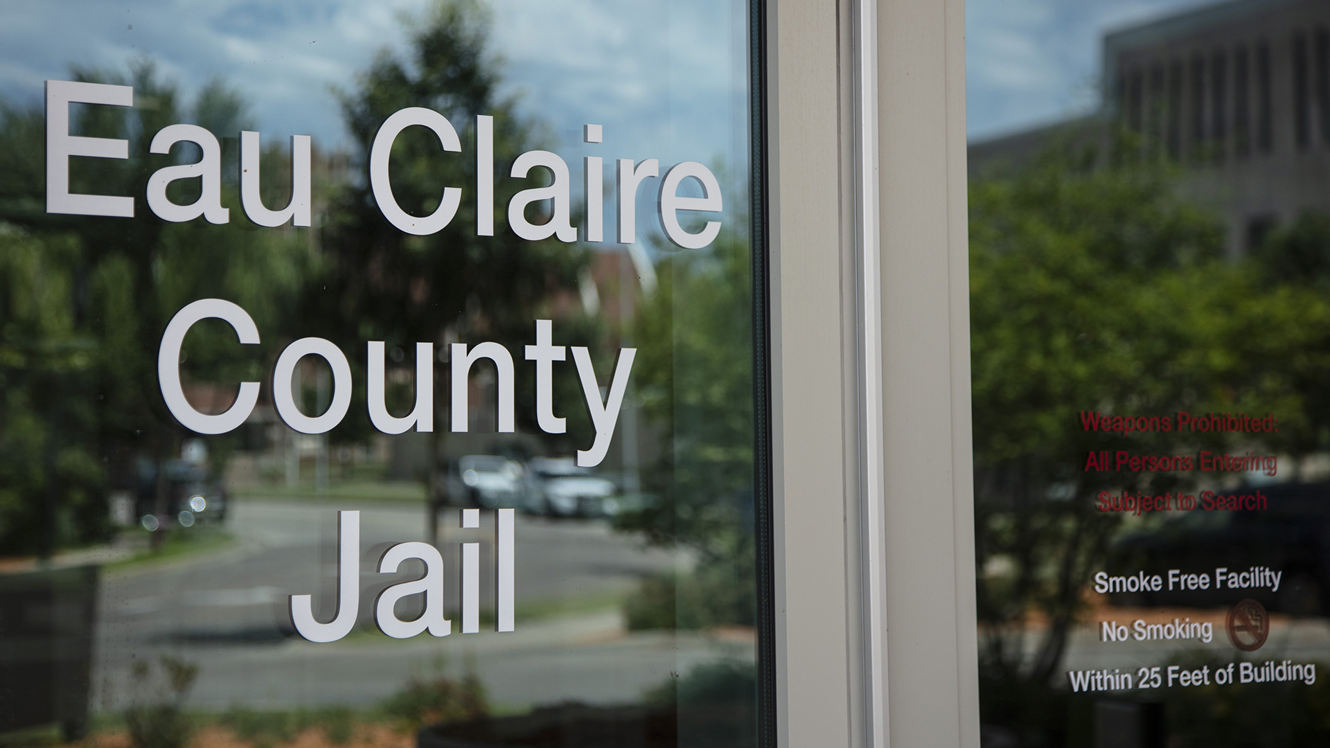 The words "Eau Claire County Jail" are stenciled on a glass pane in a door, with trees, parked cars and buildings visible in reflection.