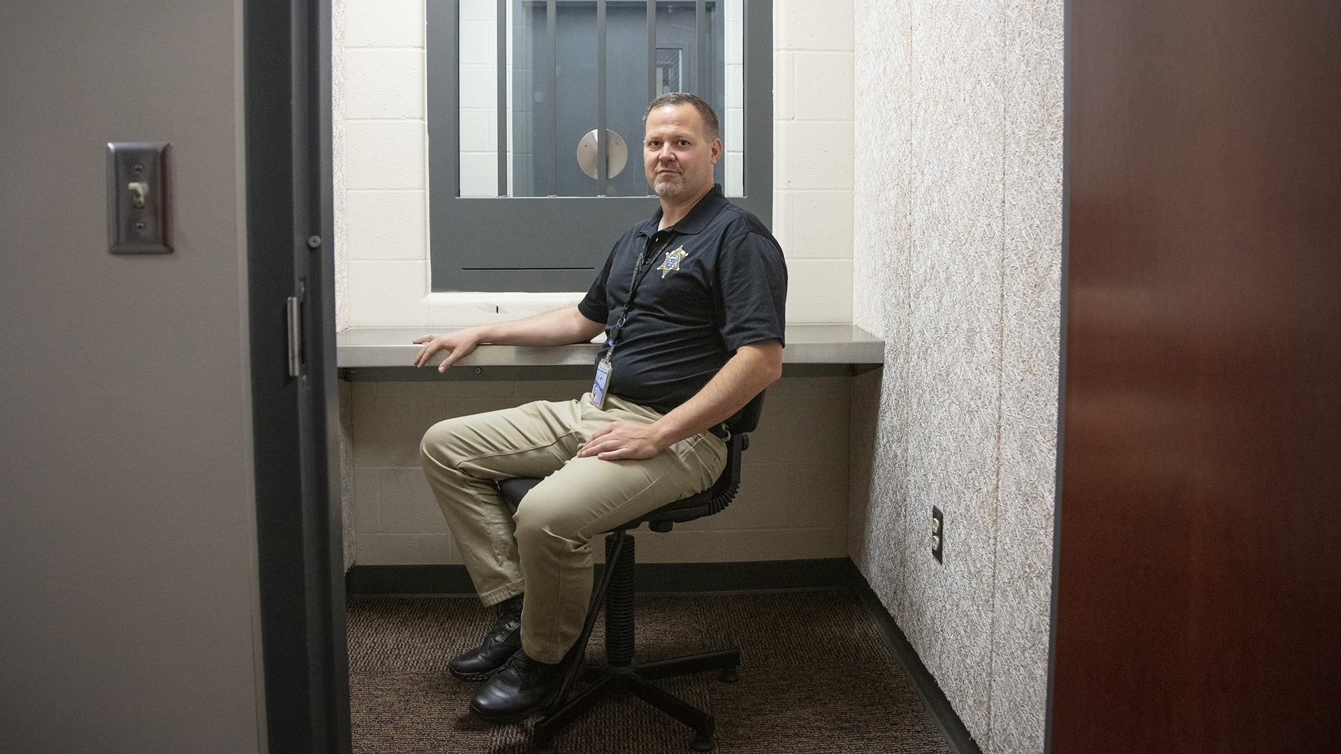 Dave Riewestahl sits in a chair inside a jail visitation room, with a connected room and door visible through a barred window.