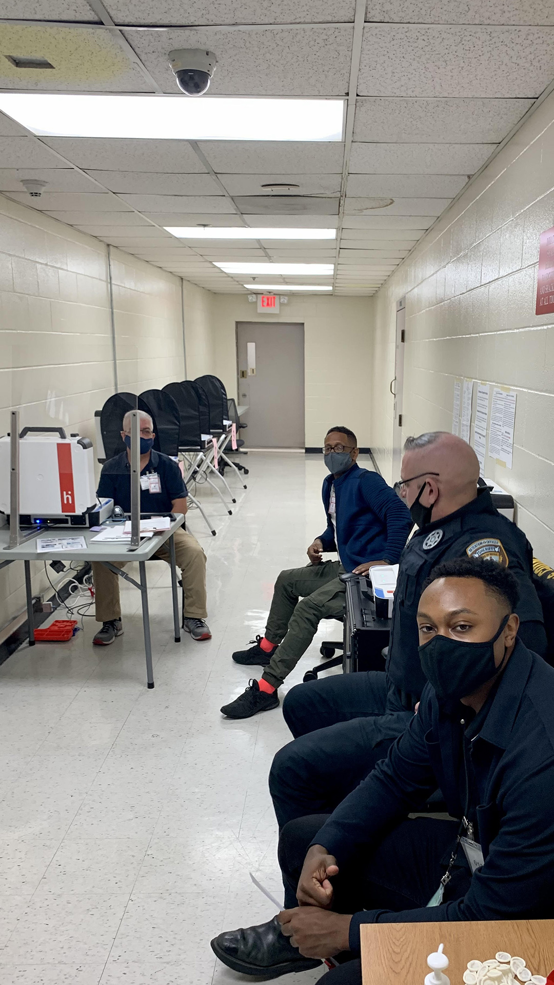 Jail staff wearing face masks sit at stations for inmate voting in a hallway with painted concrete block walls and fluorescent lights.