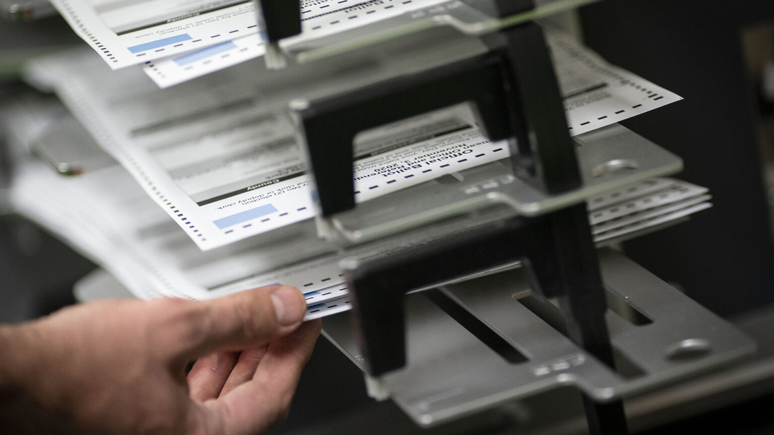 A hand grasps the corners of a ballot placed in one slot of a sorting apparatus, with other ballots in different slots above.