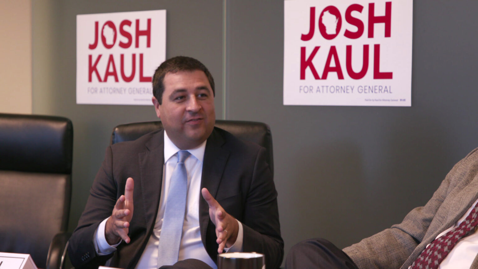 Josh Kaul sits in an office chair and gestures with both hands, with two of his campaign signs attached to the wall in the background.