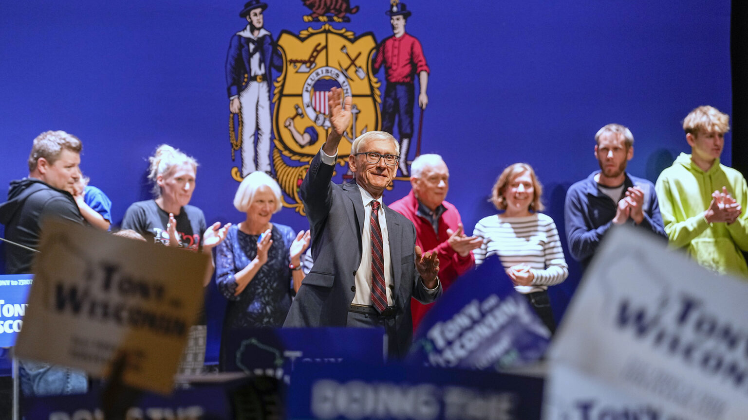 Tony Evers stands on a stage and waves with his right hand, with people standing in a line behind him clapping their hands with a large Wisconsin flag as a backdrop, while members of an audience hold up campaign signs.