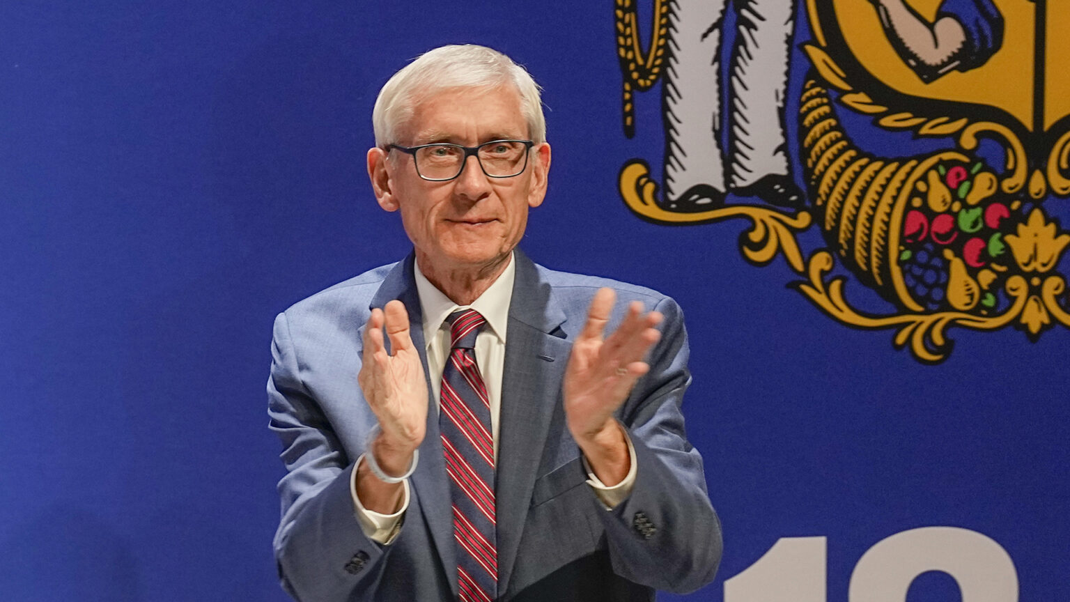 Tony Evers claps his hands while standing in front of a large image of the Wisconsin flag.