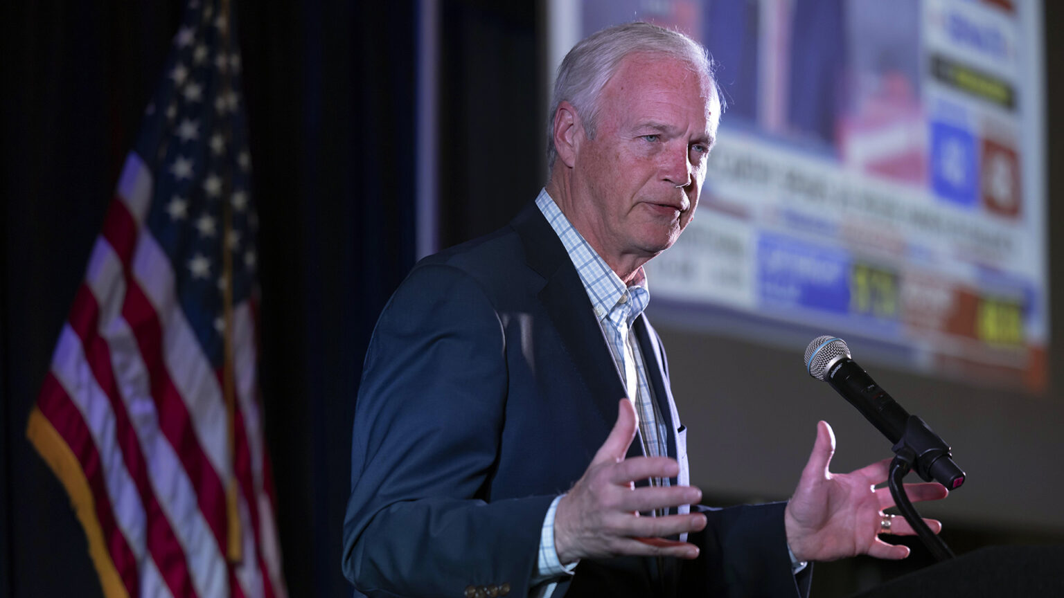 Ron Johnson stands behind a podium and speaks into a microphone while gesturing with both hands, with a U.S flags and projector screen in the background.