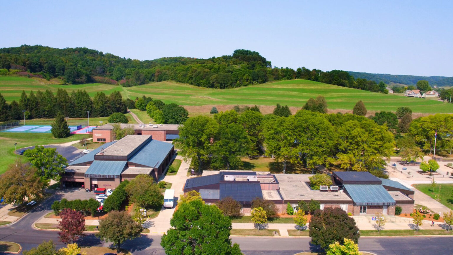 An aerial photo shows several large buildings with multiple sections situated among roads, parking lots and green areas, with hilly farm fields and woods in the background.