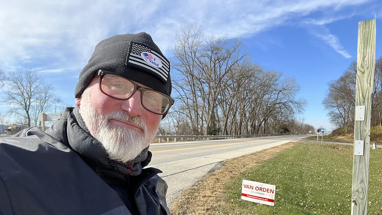 Derrick Van Orden poses for a selfie while standing next to the side of a road, with a campaign sign and trees in the background.