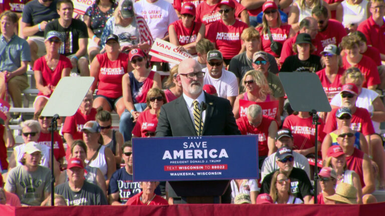 Derrick Van Orden stands behind a podium with a graphic reading Save America and speaks into a microphone while facing teleprompters, with an audience of people in the background, many wearing t-shirts for Tim Michels.
