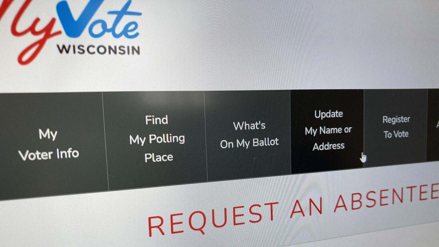 A photo shows a computer screen displaying a page on myvote.wi.gov about requesting absentee ballots, with the cursor pointing at a link titled Update My Name or Address.
