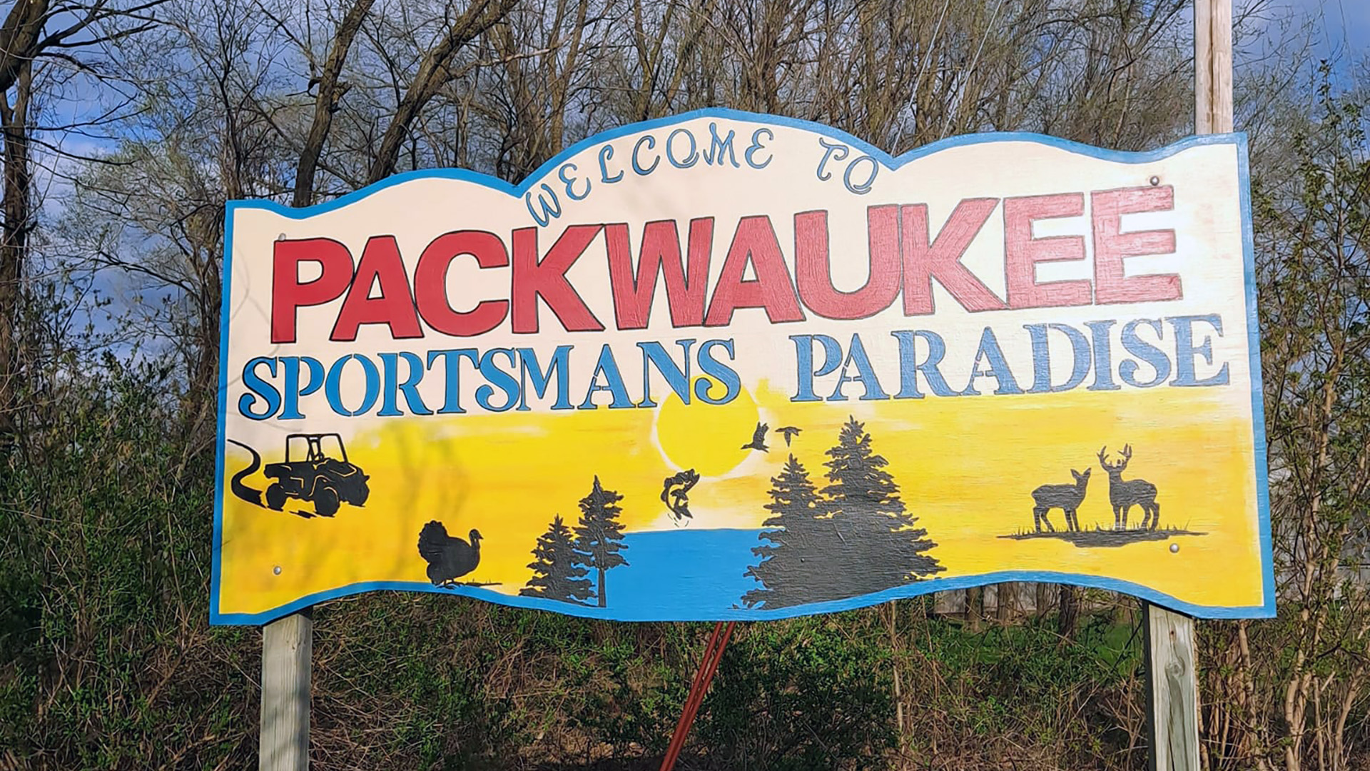 A painted wood sign reads "Welcome to Packwaukee" and "Sportsmans Paradise" above illustrations of wildlife, a UTV and trees next to water, with bushes and trees in the background.