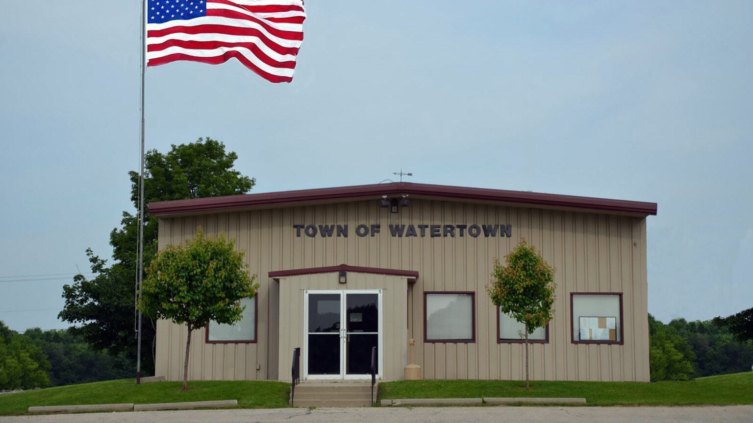 A U.S. flag flies on a flagpole next to a single-story metal-sided building with double doors, four windows and a sign reading Town of Watertown, with two small trees in front and more trees in the background.
