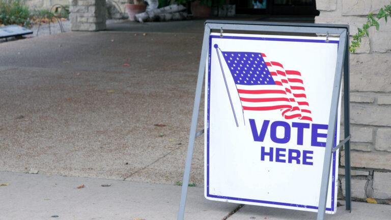 A sandwich board sign with a U.S. flag and the words Vote Here stands next to a masonry wall alongside the walkway to a building entrance.