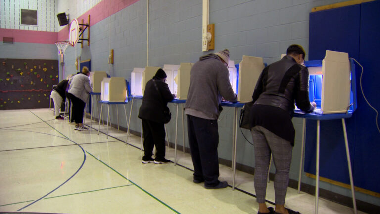 Voters stand and fill in ballots in a line of privacy booths next to a concrete wall in a gymnasium.