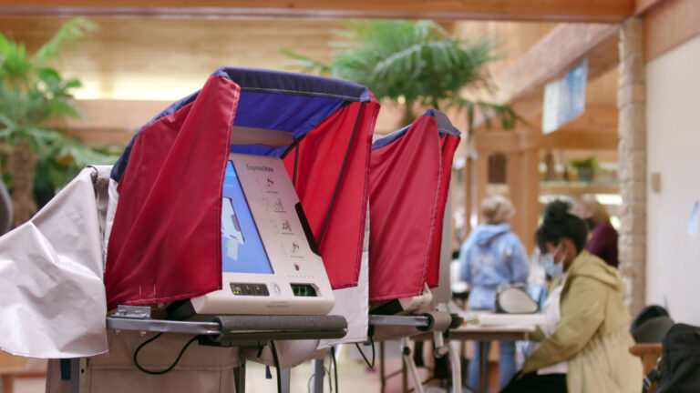 Two ballot printing machines are placed next to each other in a room, with people seated and standing in the background.