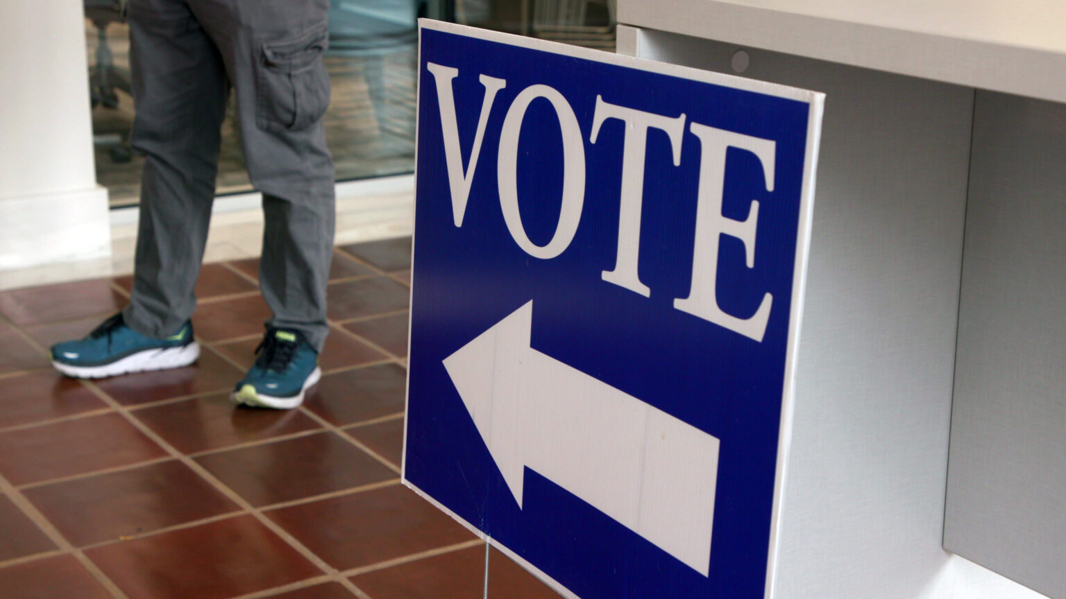 A yard sign with the word VOTE and a pointing arrow is propped against a counter in a room with tile flooring and a person standing in the background.