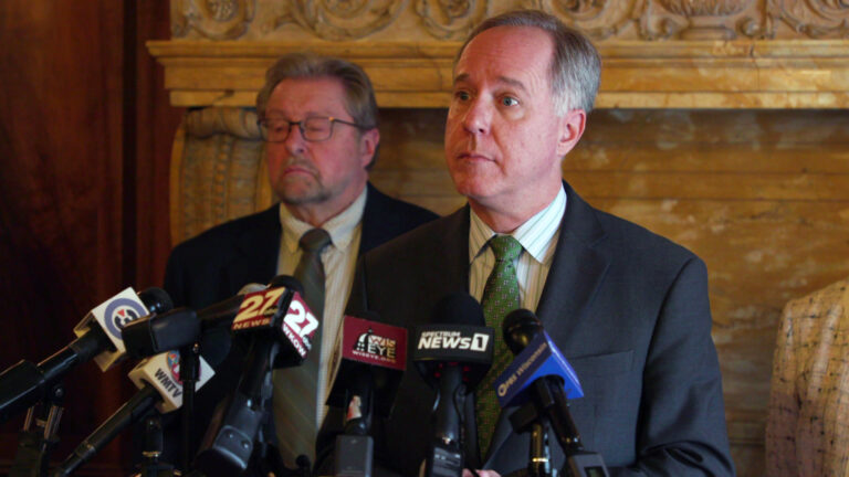 Robin Vos stands behind a podium topped with a row of microphones and listens to a question, with another person standing in the background.