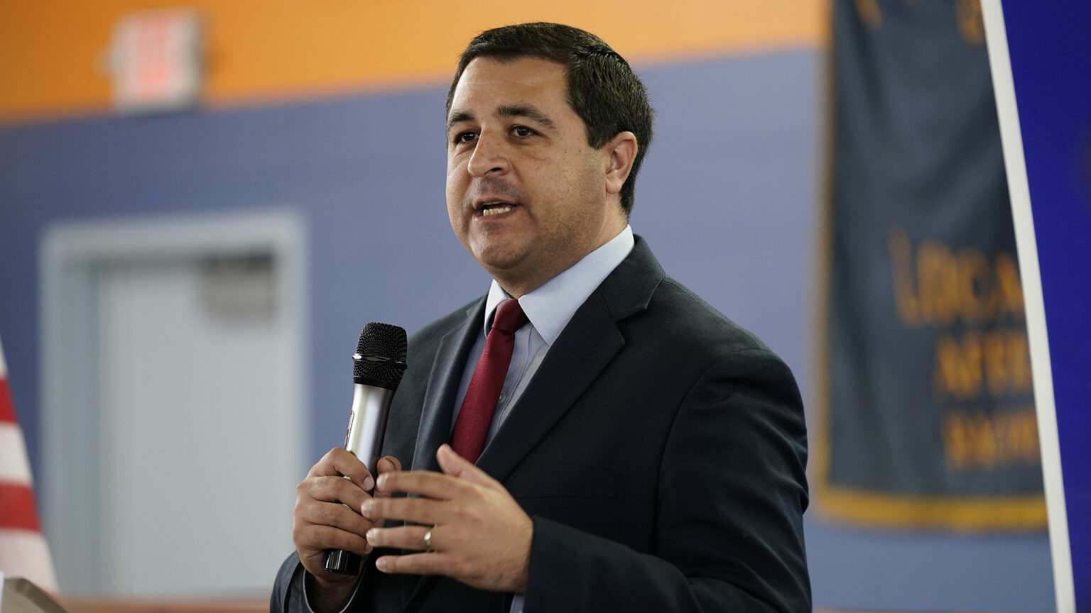 Josh Kaul stands in a room and speaks while holding a microphone in his right hand, with a door and banner in the background.