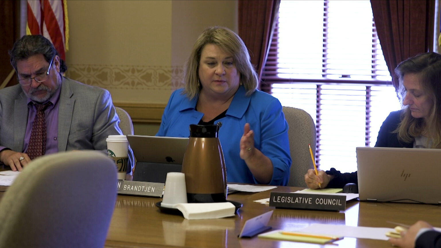 Janel Brandtjen sits at a table and gestures with her left hand while facing a laptop computer, with other people seated on either side of her and with a window and U.S. flag in the background.