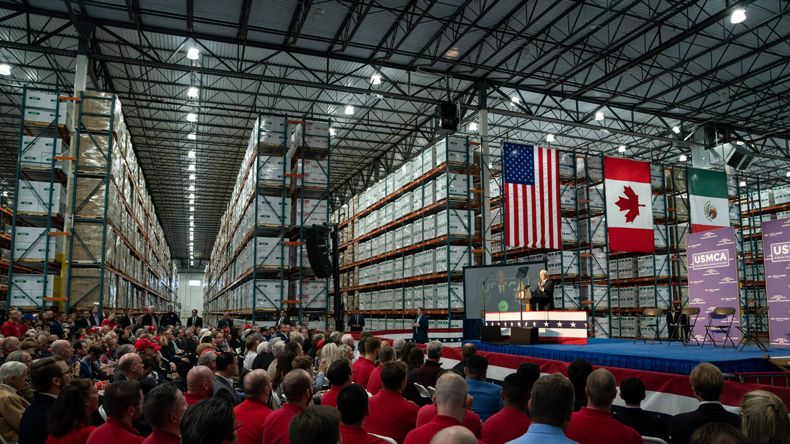 Mike Pence speaks from behind a podium on a stage in the middle of a large warehouse, facing a seated audience and with the U.S., Canada and Mexico flags mounted in front of multiple, long bays with six vertical levels of storage.
