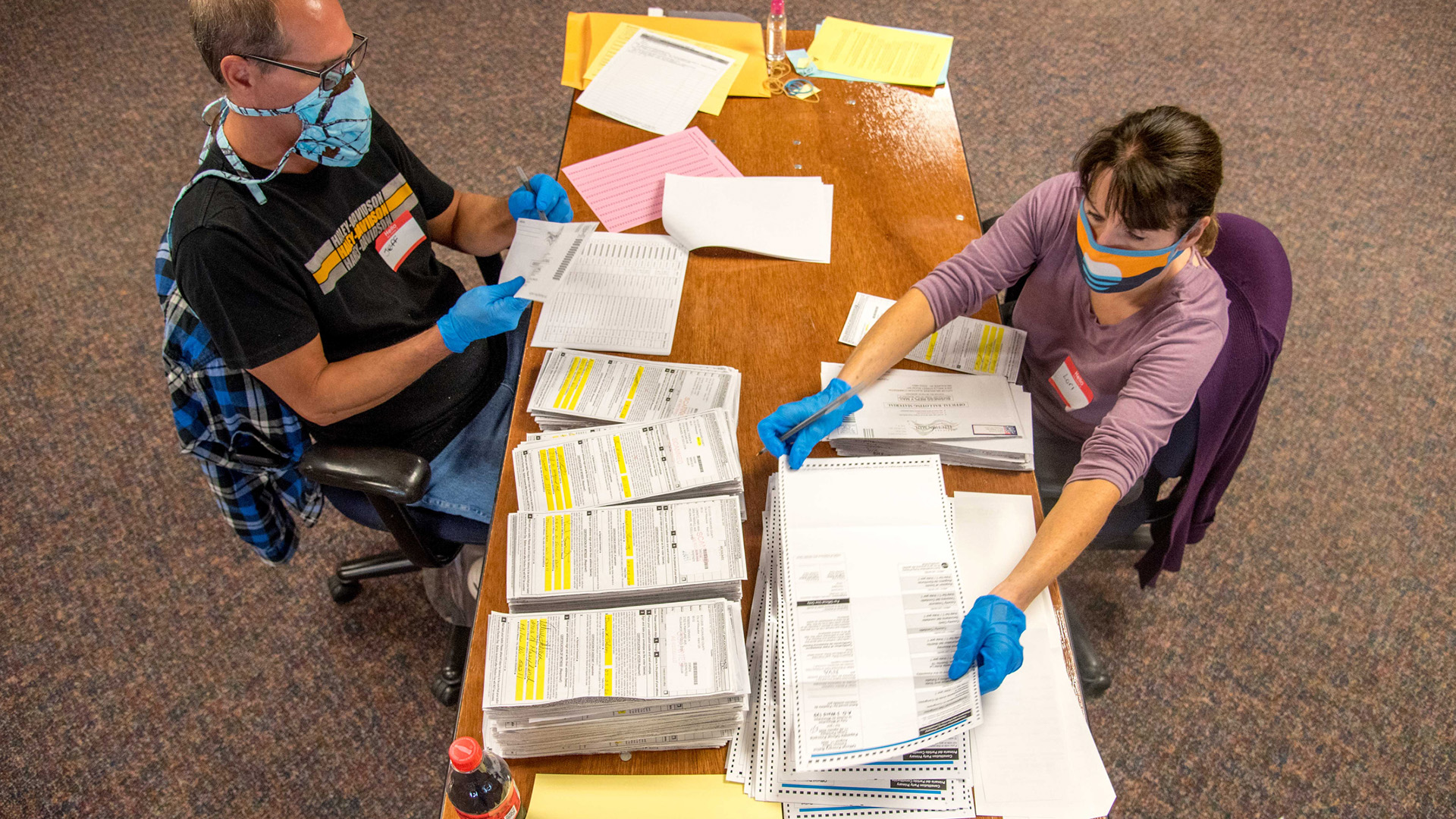 On overhead photo shows two people seated at opposite sides of a wood table covered with stacks of ballots and other election materials.