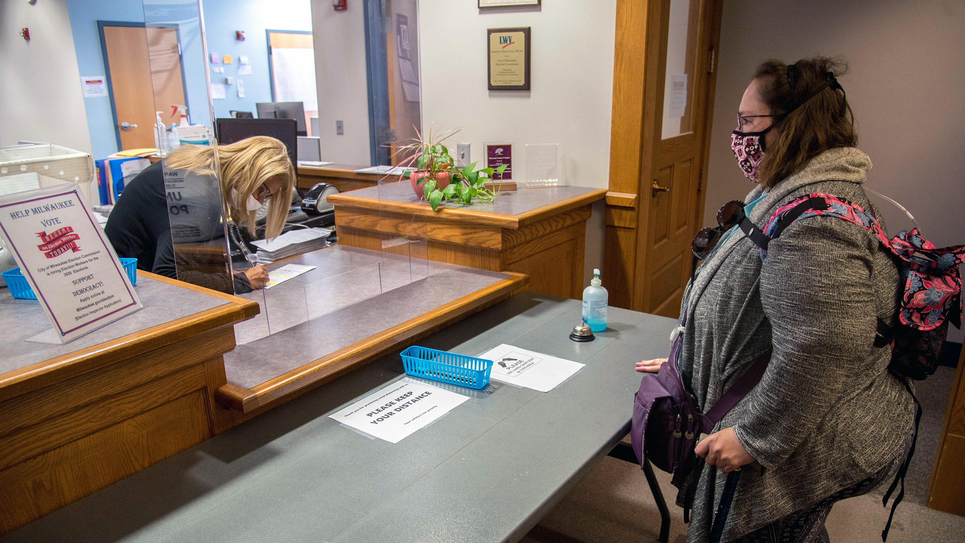 An elections worker standing behind a service counter in an office leans over and signs a ballot envelope form while Lee Ann Media watches from across a table.