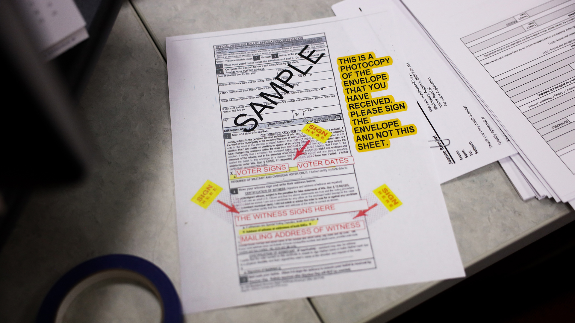 A sample ballot stamped with the word "SAMPLE" and highlighted instructions and stickers with arrows pointing toward documentation fields sits on a desk.