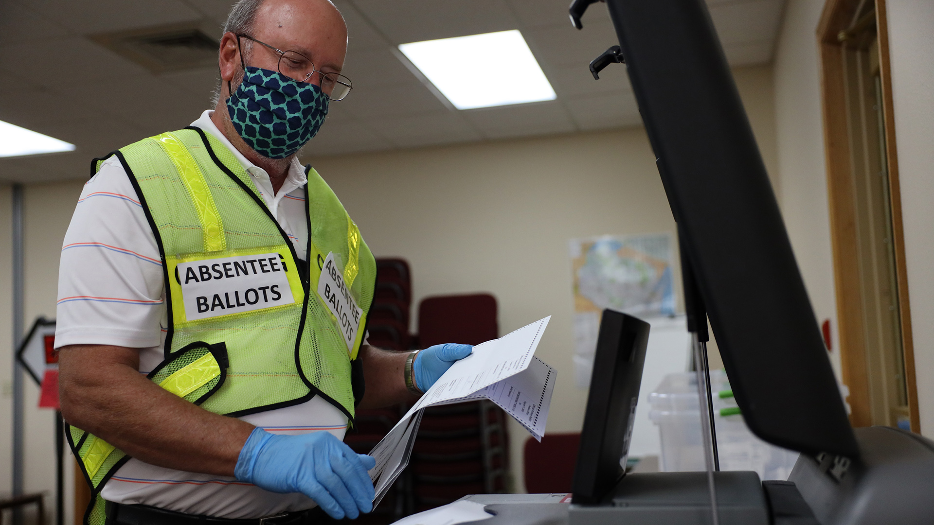 Mike Otten wears a high visibility safety vest with two labels reading "ABSENTEE BALLOTS" while holding several ballots in his left hand above a ballot tabulation machine in an office.