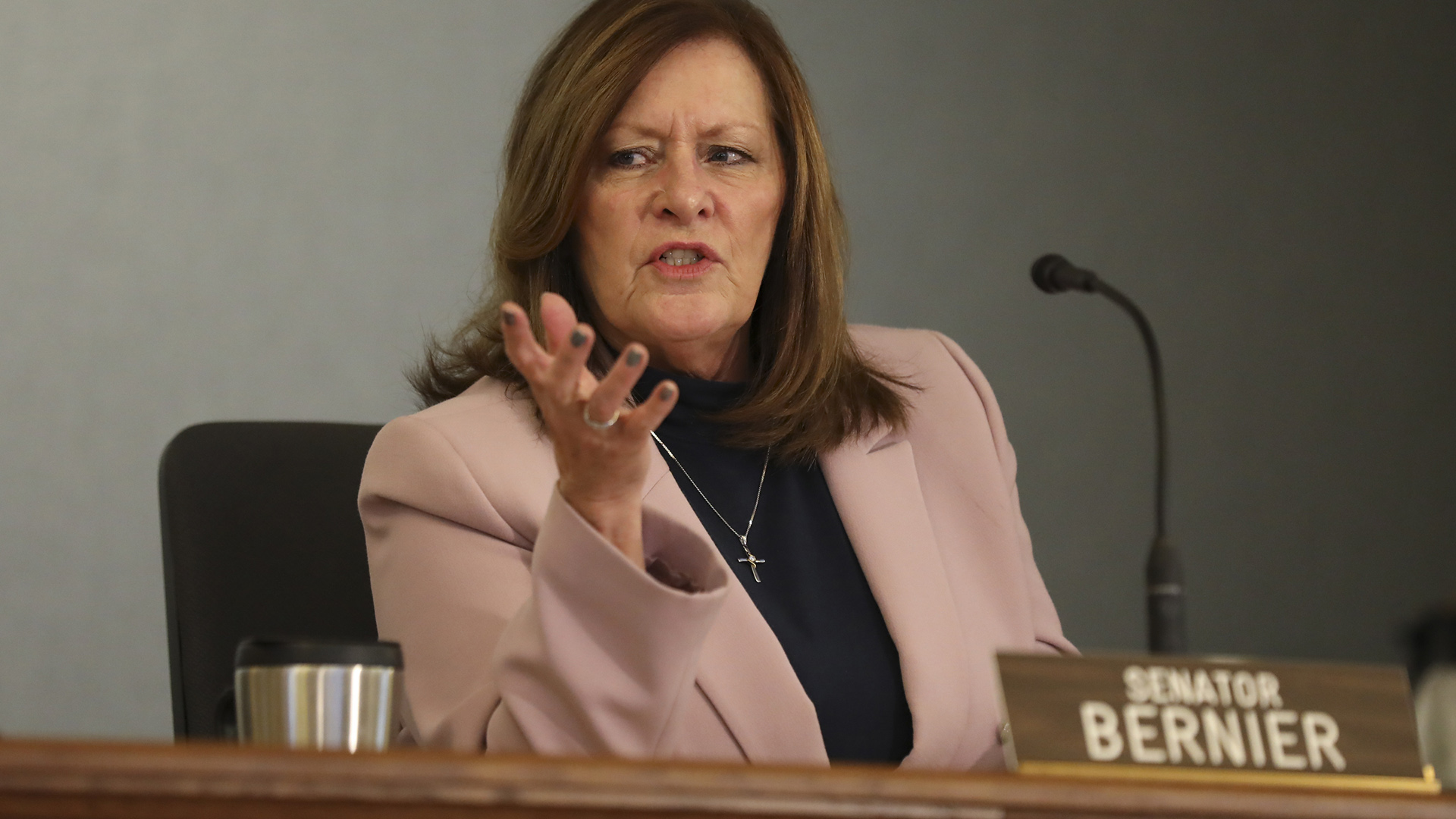 Kathleen Bernier gestures with her right hand while speaking and seated at a table outfitted with a microphone and a desk name plate reading "Senator Bernier."