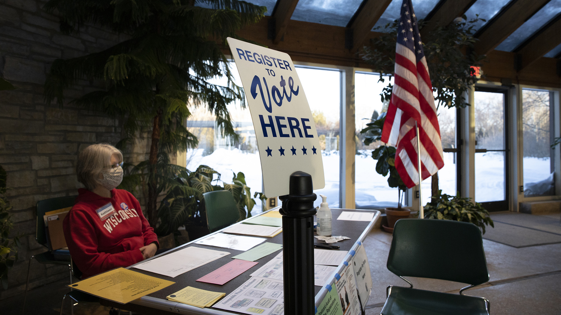 Kathleen Krchnavek sits behind a table covered with different paper forms and instructional materials, with a sign on a stand reading "Register to Vote Here" and a U.S. flag on either side, with a skylight, windows and glass doors in the background showing snowy conditions outside.