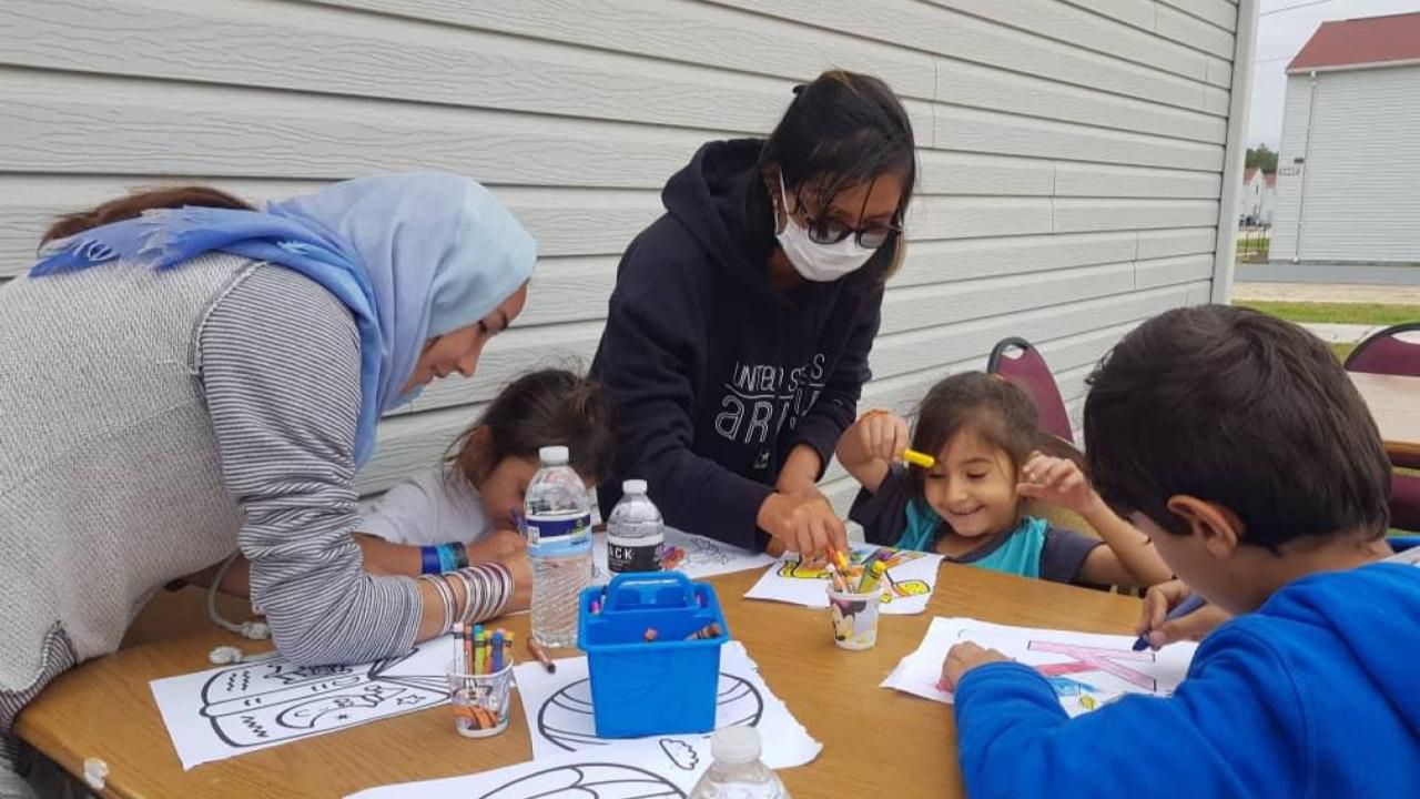 Two women stand at a table outdoors and help three seated children use crayons to illustrate coloring pages, with the siding of a building wall and other buildings in the background.