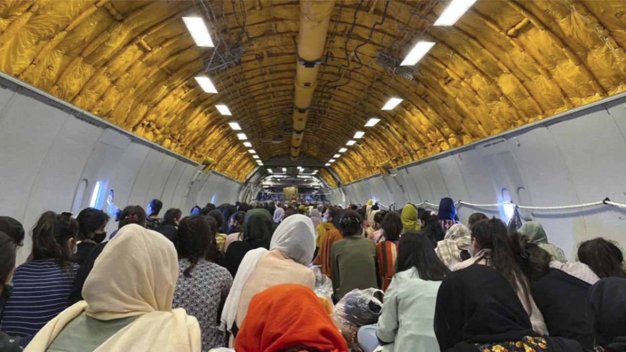 The interior fuselage of a cargo plane, outfitted with overhead lights but not other passenger infrastructure, is filled with seated people facing toward its front.
