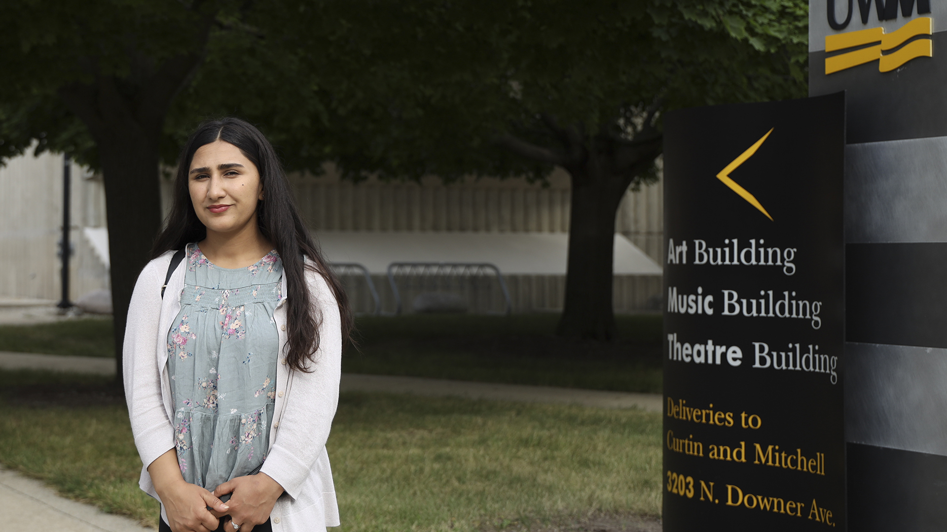 Mahrukh Delawarzad stands outdoors next to a sign with an arrow pointing toward the direction of an "Art Building," "Music Building" and "Theatre Building," with trees, bicycle racks and a building in the background.