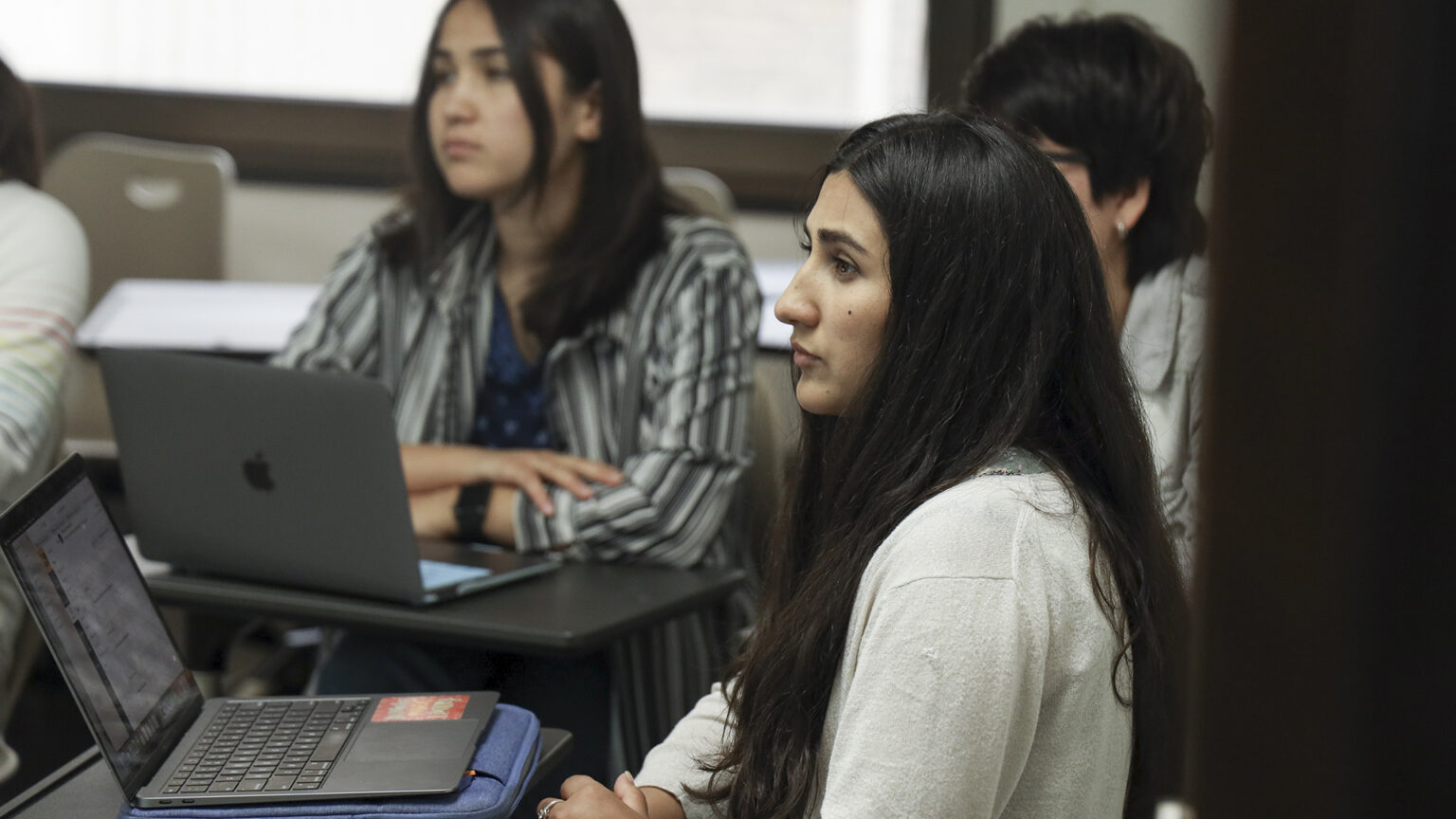 Manizha Nazari and Mahrukh Delawarzed sit in desks with open laptops in front of them in a classroom with other students.