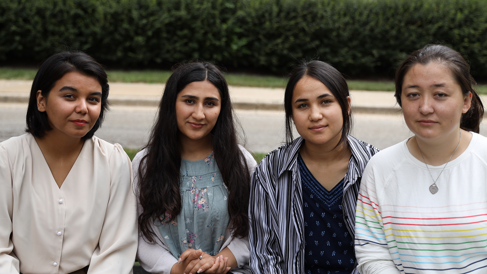 Tahera Sultani, Mahrukh Delawarzad, Manizha Nazari and Shekiba Sultani sit next to each other outdoors, with a road, sidewalk and hedge in the background.